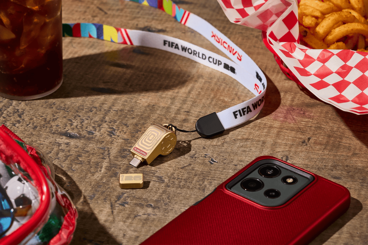 A small gold Sandisk USB drive shaped like a referee whistle lies on a wooden table. It features the FIFA World Cup emblem and comes with a matching gold cap placed nearby. Attached to the drive is a colorful lanyard with “FIFA WORLD CUP” printed on a white strap. Surrounding the USB drive are several items: a red‑cased smartphone, a basket of curly fries in red‑and‑white checkered paper, a cup of iced soda, and part of a clear pouch filled with objects. The scene has a casual, outdoor, game‑day feel.