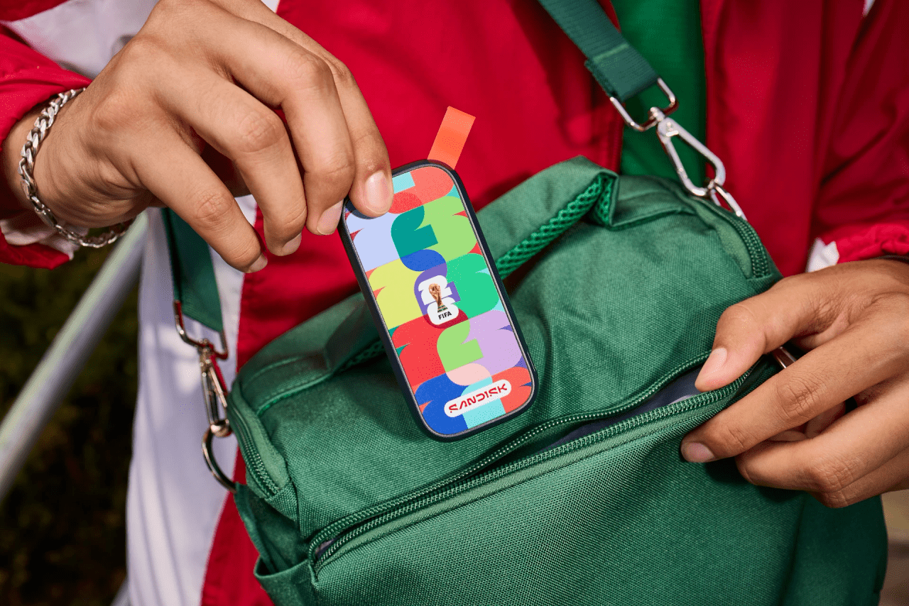 A person is holding a colorful Sandisk portable SSD decorated with a bright, abstract pattern in red, green, yellow, and blue. The drive features an official FIFA World Cup emblem in the center. The person is placing or removing the SSD from the front pocket of a green bag. They are wearing a red and white jacket, and only their hands and part of the bag are visible.